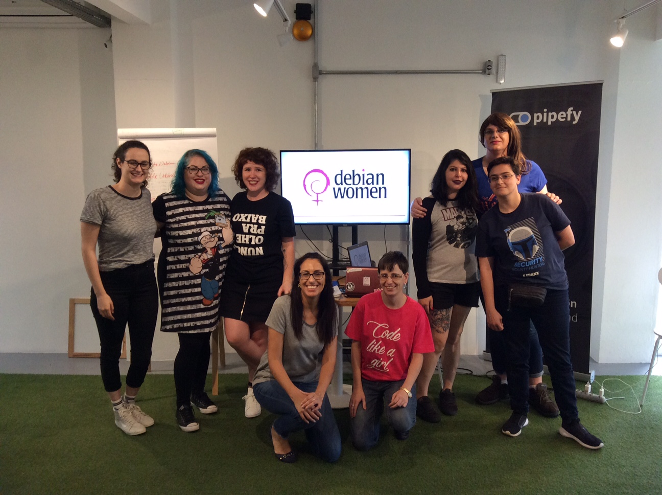 The eight women who attended the meeting gathered together in front of a tv with the Debian Women logo