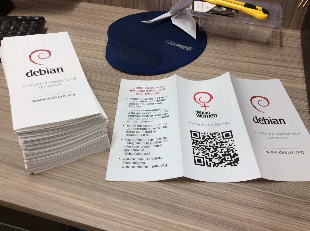 A pile with folded Debian Women folders. The writings are in Portuguese and it's possible to see a QR code.