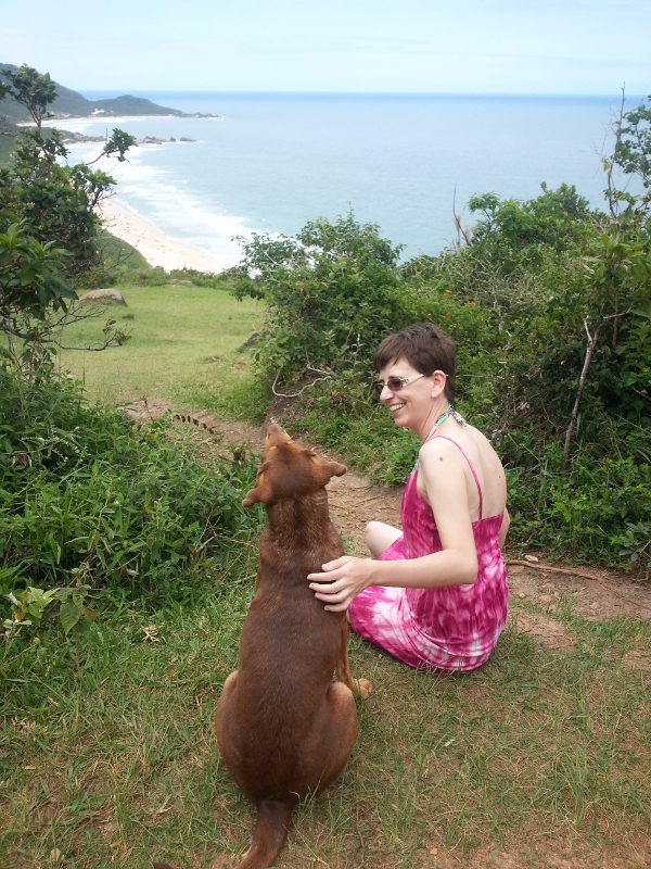 Renata's picture, a white woman sitting on the grass, overlooking the beach below. She pets a brown stray that sits next to her